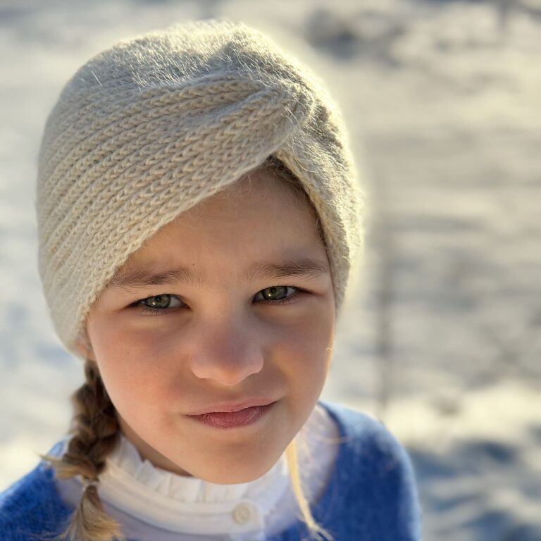 Jente i lys beige Vinterturban med vridd front, med flette og blå cardigan